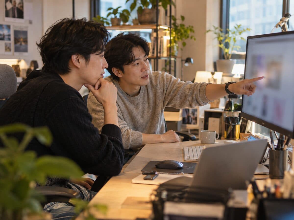 작은 IT 사무실에서 개발자 두 명이 모니터를 보며 홈페이지 시안을 검토하는 모습